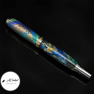 May include: A handcrafted fountain pen with a vibrant design. The pen features a mix of blue, green, and gold hues, with intricate patterns and metallic accents. The pen's tip and end are silver-colored. The pen is labeled "ACinked Functional Art".