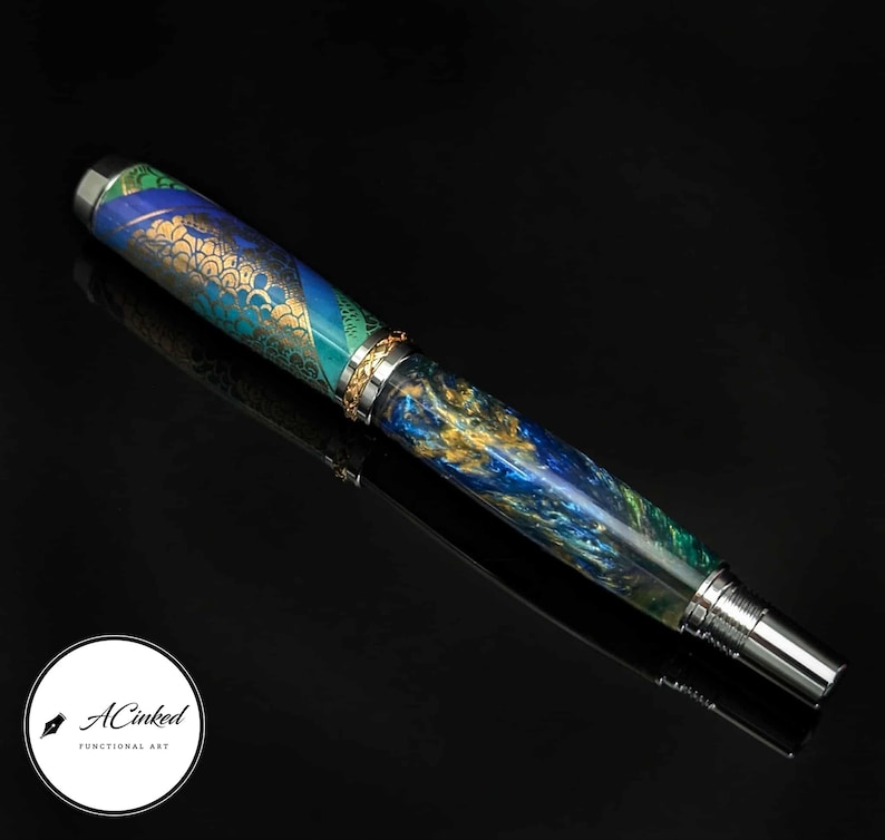 May include: A handcrafted fountain pen with a vibrant design. The pen features a mix of blue, green, and gold hues, with intricate patterns and metallic accents. The pen's tip and end are silver-colored. The pen is labeled "ACinked Functional Art".