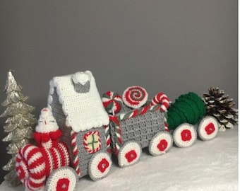 May include: A crocheted Christmas train with a white roof, gray body, and red and white striped accents. The train features round wheels with red centers and is decorated with candy and a green tree. A silver Christmas tree and pine cone are also present.
