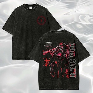 May include: Two-sided black t-shirt with a distressed wash. The front features a red emblem, while the back displays a red graphic of a character and the words "DOOM SLAYER" in a vertical font. The shirt has short sleeves and a crew neck.