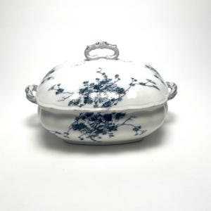 May include: A white porcelain serving dish with a lid, adorned with a blue floral pattern. The dish has a rounded shape with handles on the sides and a decorative handle on the lid. A classic piece for serving food.