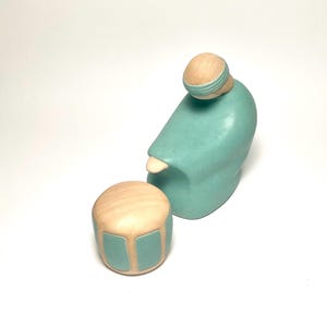 May include: A teal ceramic sculpture of a seated figure with a light wood-colored head and a matching small drum. The figure is in a seated position, with the head slightly tilted. The drum has teal accents.
