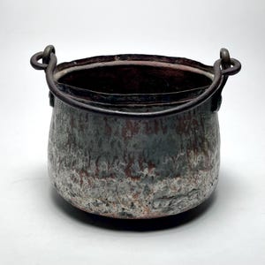May include: A small, textured metal cauldron with a dark, weathered appearance. The pot has a rounded body, a wide opening, and a sturdy handle. The exterior features a mix of gray, brown, and white tones, suggesting age and use. The handle is made of dark metal.