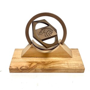 May include: A bronze-colored geometric sculpture featuring a square within a circle, mounted on a wooden base. The square has a stylized design. The sculpture is a decorative art piece.