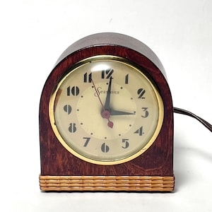 May include: A vintage Sessions clock with a dark wood case and a gold-tone bezel. The clock face is cream-colored with black numerals and hands. The clock has a woven base and a power cord.