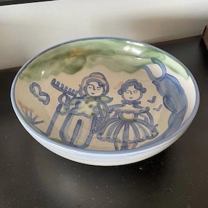 May include: A ceramic serving bowl with a hand-painted design. The bowl features a light blue rim and a scene with two figures in blue and green, with a green and blue background.