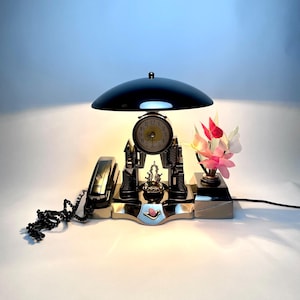 May include: A vintage desk lamp with a black shade, a clock, and a telephone. The clock has a gold face and is surrounded by a cityscape design. A small floral arrangement and a telephone are also present.