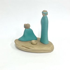 May include: A ceramic nativity scene featuring two figures in teal robes and a small bowl, set on a beige base. One figure stands, the other kneels. The figures have tan-colored head coverings.