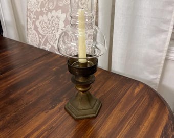 May include: A vintage brass candlestick with a clear glass hurricane shade. The candleholder has a hexagonal base and a tiered design. A lit, cream-colored candle is inside the glass shade. The candlestick sits on a dark wooden table.