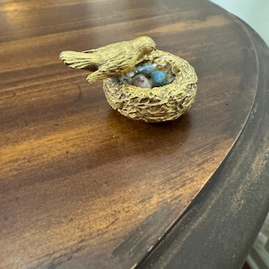 May include: A gold-colored bird figurine perched above a nest with blue eggs. The nest is intricately detailed, and the bird has textured feathers. The figurine is placed on a wooden surface.