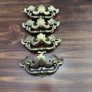 Könnte beinhalten: Vier antike Schubladengriffe aus Messing mit aufwendigen Designs. Jeder Griff hat einen gebogenen Griff und eine dekorative Rückplatte. Die Griffe sind auf einer dunklen Holzoberfläche angeordnet und zeigen ihre Vintage-Ästhetik.