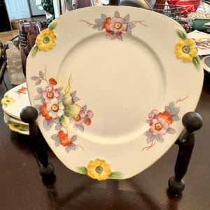 May include: A set of four white ceramic plates with a floral design. The plates have a scalloped edge and are decorated with yellow, pink, and orange flowers. The plates are displayed on a black metal stand.