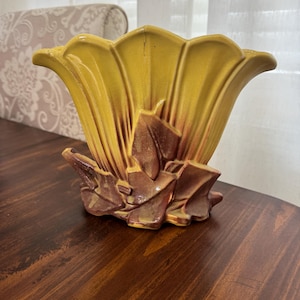 Puede incluir: Un jarrón de cerámica amarillo y marrón con un diseño floral. El jarrón tiene una parte superior acampanada en forma de pétalo y una base texturizada y en capas. El jarrón está sobre una superficie de madera oscura.