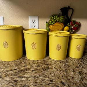 Peut inclure: Un ensemble de quatre boîtes de rangement cylindriques jaune vif avec des couvercles assortis. Chaque boîte présente un motif floral en forme d'étoile marron centré sur le devant. Les contenants varient en taille, du plus grand au plus petit.