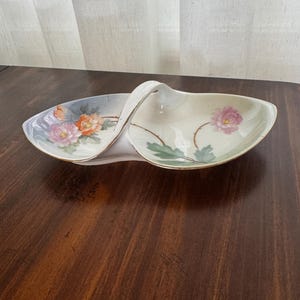 May include: A two-sectioned porcelain serving dish with a central arched handle. The dish features a cream-colored base with hand-painted floral motifs in shades of pink, orange, and green, accented by a delicate gold trim along the edges.