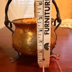 May include: A copper pot with a rounded body, three metal feet, and a twisted metal handle. A white ruler is positioned vertically against the pot, showing a height of approximately 6 inches from the base to the top rim of the vessel.