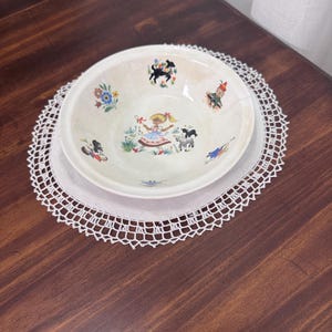 May include: A vintage ceramic bowl with a whimsical illustration of a girl, animals, and flowers. The bowl is set on a white lace doily, resting on a dark wooden surface. The bowl's design features a light beige background with colorful illustrations.