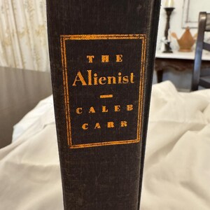 Op de afbeelding: Een donkerbruin hardback boek met gouden letters op de rug. De titel is "The Alienist" van Caleb Carr.