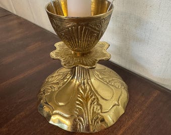 May include: A gold-colored candle holder with ornate floral and shell-like designs. The candle holder has a wide, decorative base and a cup-shaped top to hold a white candle. Two metal clips are holding the candle in place.