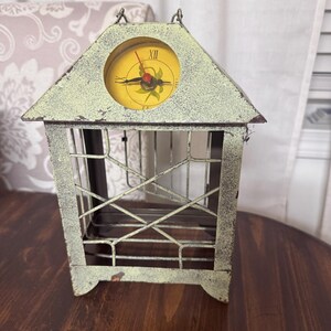 May include: A decorative, vintage-style clock in a cage-like design. The clock face is yellow with black hands and Roman numerals. The cage is made of metal with a distressed, off-white finish, and sits on a wooden surface.