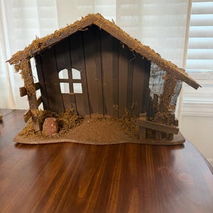 May include: A brown wooden stable structure featuring a gabled roof covered in dried moss, vertical wall paneling, a single square window cutout, a small fence section on the right, and a textured ground base with scattered moss and a small stone.