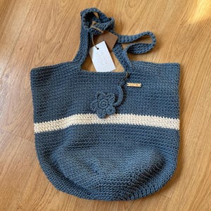 May include: A blue crocheted tote bag with a white stripe and a crocheted flower. The bag has a wooden tag that says "hecho a mano".