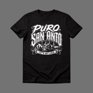 May include: Black t-shirt with a white graphic design. The design features the words "PURO SAN ANTONIO" in a stylized font, along with an illustration of the city skyline and the phrase "LET'S GET LOUD".