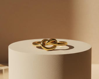 May include: A gold ring featuring a knot design. The ring is presented on a white cylindrical plinth against a neutral backdrop. The knot design is the central element, highlighting its intricate details and polished finish.