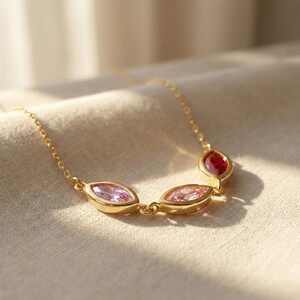 Könnte beinhalten: Eine goldfarbene Halskette mit drei ovalen Edelsteinen in Rosa- und Rottönen. Die Edelsteine sind in goldenen Fassungen gefasst und durch eine filigrane Goldkette verbunden. Die Halskette wird auf einem neutralfarbenen Stoff präsentiert.