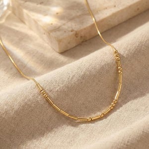 May include: A delicate gold-colored necklace with a series of small, cylindrical and spherical beads. The necklace is displayed on a beige fabric surface, with a neutral background. The jewelry is simple and elegant.
