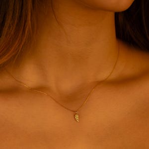 Puede incluir: Un delicado collar de oro con un pequeño colgante de hoja. El collar lo lleva una persona de tez clara.
