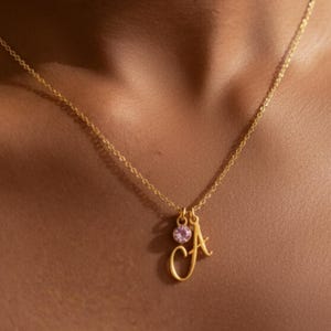 Könnte beinhalten: Eine goldfarbene Halskette mit einem Anhänger mit dem Buchstaben "A" in Schreibschrift, begleitet von einem kleinen, runden, rosa Edelstein. Die Halskette wird vor einem hautfarbenen Hintergrund gezeigt.