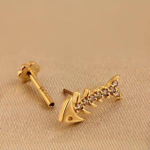 Pode incluir: Brinco dourado em forma de espinha de peixe com pequenas pedras preciosas transparentes. Mostrado ao lado de um pino dourado com uma parte superior em forma de flor. Projetado para piercing.