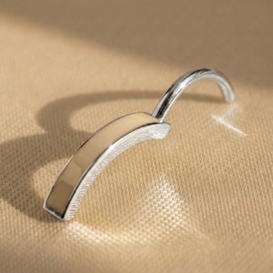 Puede incluir: Un cierre de joyería de metal plateado con una barra curva rellena de esmalte beige y un asa curva. El cierre se encuentra sobre un fondo neutro y texturizado, resaltando su elegante diseño.