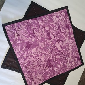 Könnte beinhalten: Zwei quadratische Topflappen aus Stoff mit einem Marmormuster in hellen und dunklen Violetttönen. Jedes Stück ist mit einem einfarbigen, dunkelbraunen Rand und diagonalen Nähten auf der Oberfläche versehen, was eine gesteppte Struktur ergibt.