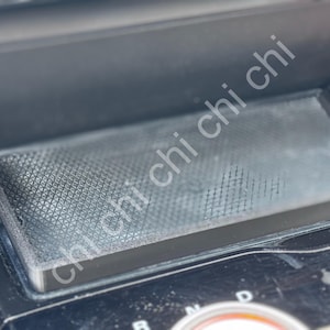May include: A close-up of a car's interior, showing a rectangular, textured, dark grey tray. The tray is set into a black dashboard, with the gear shift indicator visible below. The image is well-lit, highlighting the details of the car's interior.