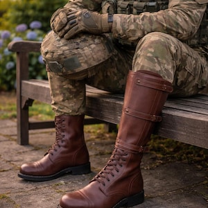 May include: Brown leather knee-high boots with laces and buckles. The boots are worn by a person in camouflage clothing, sitting on a wooden bench. The boots have a classic design and are made of leather.