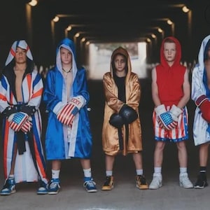 Puede incluir: Cinco personas vestidas de boxeadores están de pie en un túnel. Llevan guantes de boxeo y batas en rojo, blanco, azul y dorado. Los guantes y los pantalones cortos presentan diseños de la bandera estadounidense. Los boxeadores llevan zapatillas.