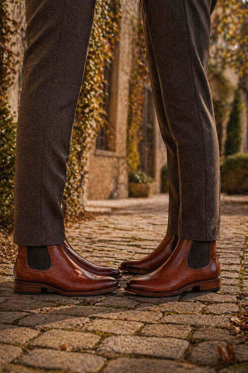 Puede incluir: Dos pares de botines Chelsea de cuero marr&oacute;n con paneles laterales el&aacute;sticos. Las botas se usan con pantalones marrones oscuros. La imagen est&aacute; tomada al aire libre en un camino adoquinado, con un fondo borroso.