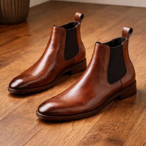 Puede incluir: Un par de botines Chelsea de cuero marr&oacute;n con paneles laterales el&aacute;sticos. Los botines tienen un acabado pulido y se exhiben sobre un suelo de madera. Los botines tienen una leng&uuml;eta en la parte trasera.