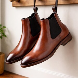 Puede incluir: Un par de botines Chelsea de cuero marr&oacute;n colgados de un perchero de madera. Los botines tienen paneles laterales el&aacute;sticos y un tac&oacute;n bajo. El cuero tiene un acabado pulido y un c&aacute;lido tono marr&oacute;n.