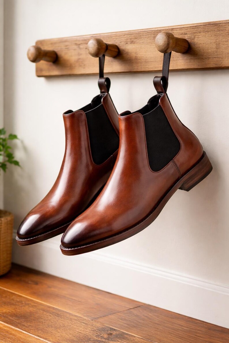 Puede incluir: Un par de botines Chelsea de cuero marr&oacute;n colgados de un perchero de madera. Los botines tienen paneles laterales el&aacute;sticos y un tac&oacute;n bajo. El cuero tiene un acabado pulido y un c&aacute;lido tono marr&oacute;n.