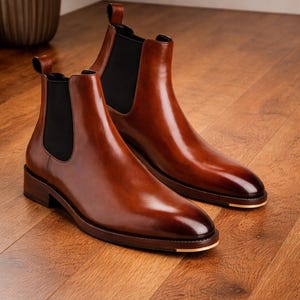 Puede incluir: Un par de botines Chelsea de cuero marr&oacute;n pulido. Los botines tienen paneles laterales el&aacute;sticos y una leng&uuml;eta. Las suelas son de color marr&oacute;n oscuro con un acento dorado en la puntera. Los botines est&aacute;n sobre un suelo de madera.
