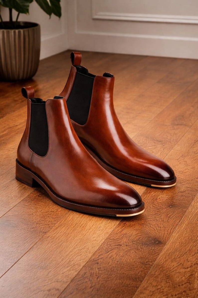 Puede incluir: Un par de botines Chelsea de cuero marr&oacute;n pulido. Los botines tienen paneles laterales el&aacute;sticos y una leng&uuml;eta. Las suelas son de color marr&oacute;n oscuro con un acento dorado en la puntera. Los botines est&aacute;n sobre un suelo de madera.