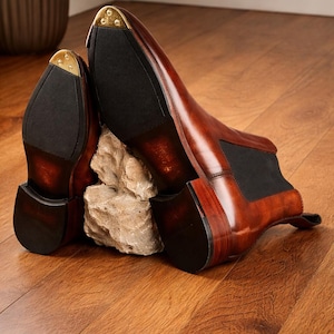 Puede incluir: Un par de botines Chelsea de cuero marr&oacute;n con paneles laterales el&aacute;sticos negros y punteras de tal&oacute;n de metal dorado. Los botines se exhiben sobre un suelo de madera, mostrando su acabado pulido y dise&ntilde;o cl&aacute;sico. Talla 43.