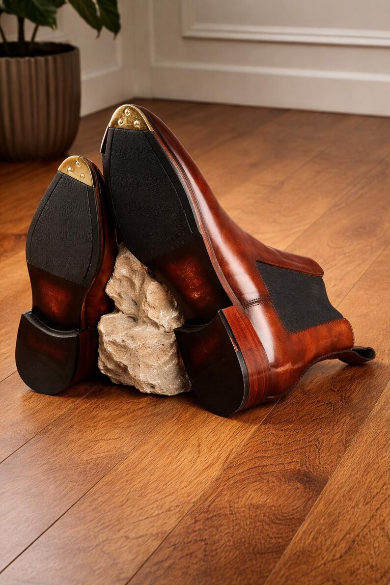 Puede incluir: Un par de botines Chelsea de cuero marr&oacute;n con paneles laterales el&aacute;sticos negros y punteras de tal&oacute;n de metal dorado. Los botines se exhiben sobre un suelo de madera, mostrando su acabado pulido y dise&ntilde;o cl&aacute;sico. Talla 43.