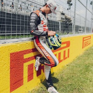 Puede incluir: Un piloto de carreras con un traje de carreras Haas, apoyado en una pared amarilla y roja con el logo de Pirelli. El piloto sostiene un casco con detalles azules y amarillos. La escena es al aire libre en un d&iacute;a soleado.
