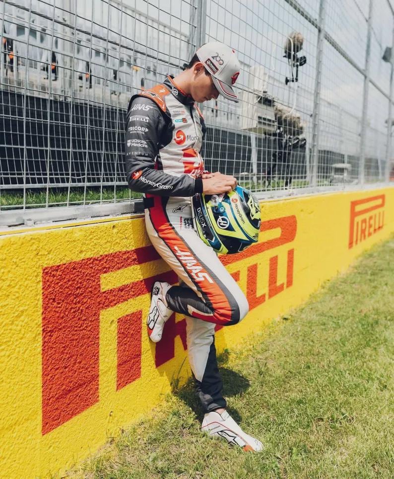 Puede incluir: Un piloto de carreras con un traje de carreras Haas, apoyado en una pared amarilla y roja con el logo de Pirelli. El piloto sostiene un casco con detalles azules y amarillos. La escena es al aire libre en un d&iacute;a soleado.