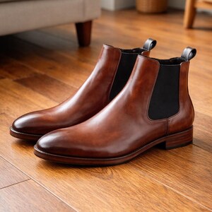 Puede incluir: Un par de botines Chelsea de cuero marr&oacute;n con paneles laterales el&aacute;sticos. Las botas tienen un acabado pulido y est&aacute;n sobre un suelo de madera. Las botas son de estilo cl&aacute;sico.