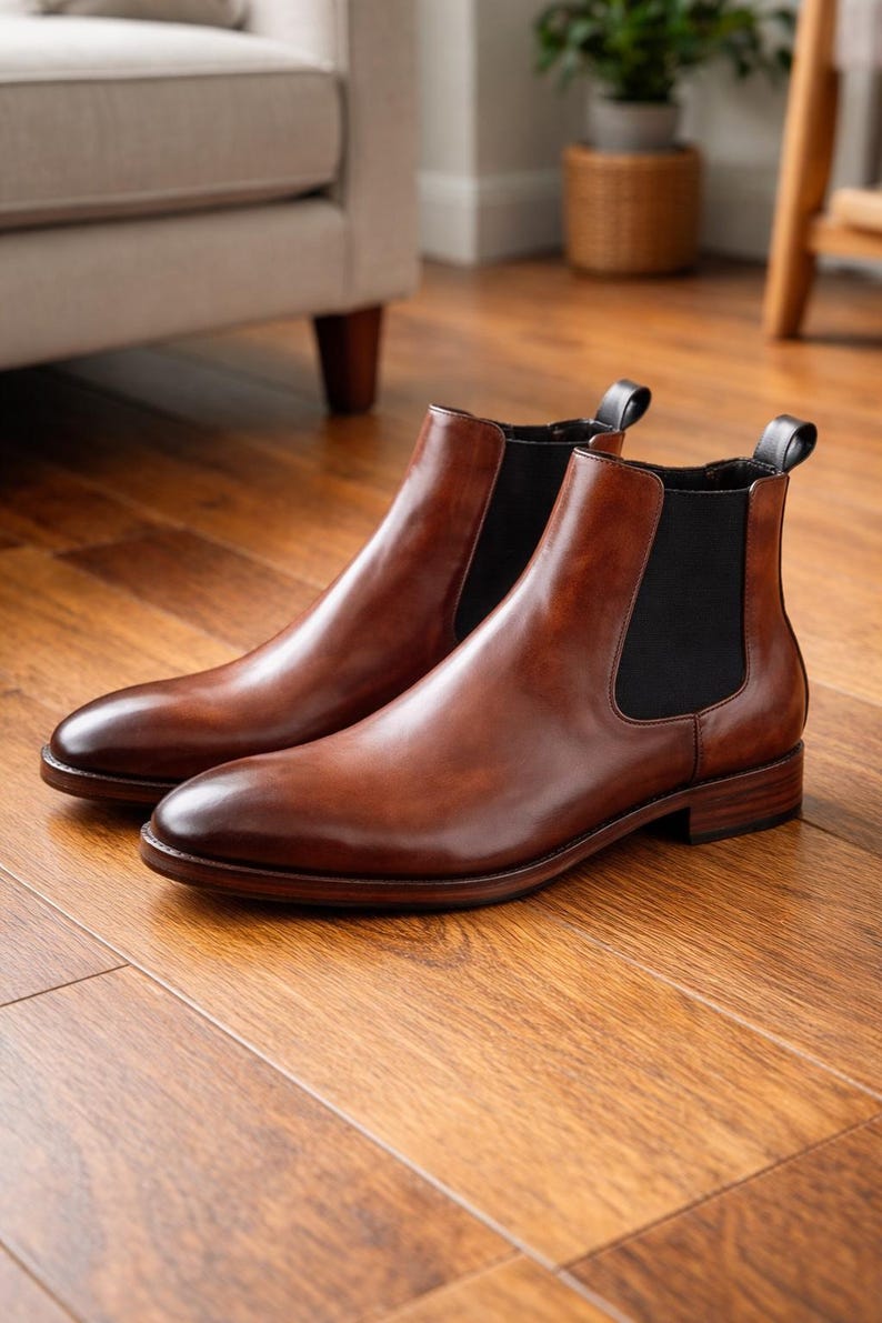 Puede incluir: Un par de botines Chelsea de cuero marr&oacute;n con paneles laterales el&aacute;sticos. Las botas tienen un acabado pulido y est&aacute;n sobre un suelo de madera. Las botas son de estilo cl&aacute;sico.